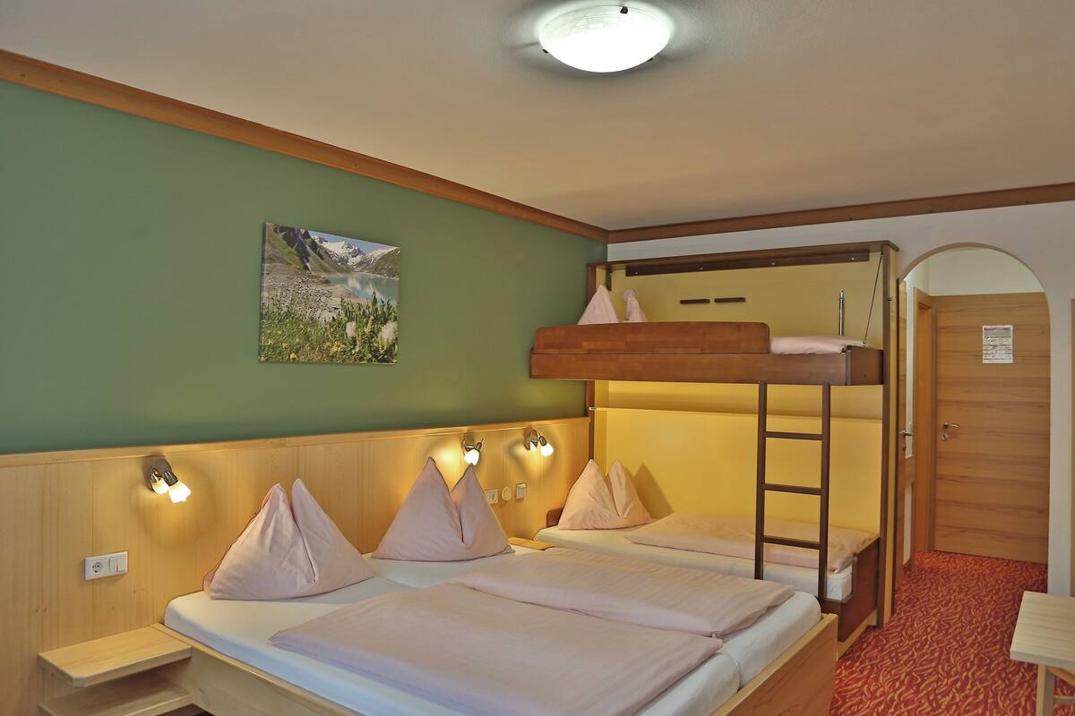 Habitación de hotel con dos camas, una litera, una mesita pequeña, un banco y un cuadro en la pared.
