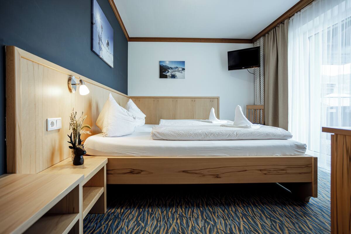 Habitación de hotel con cama de madera, mesita de noche con lámpara, televisor montado en la pared y ventana con cortinas.