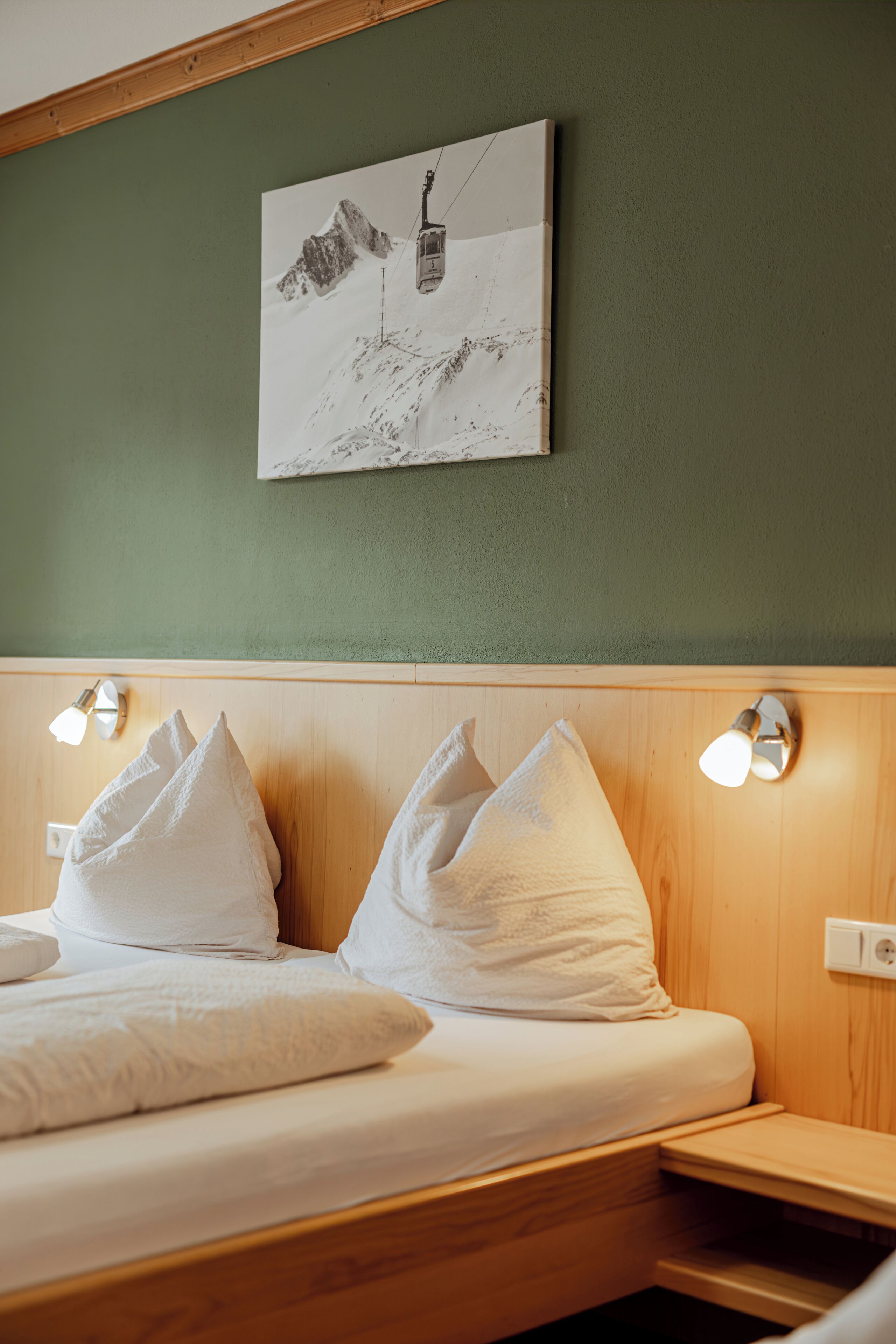A hotel room with a bed, pillows, a headboard with wall lights, and a painting of a mountain.
