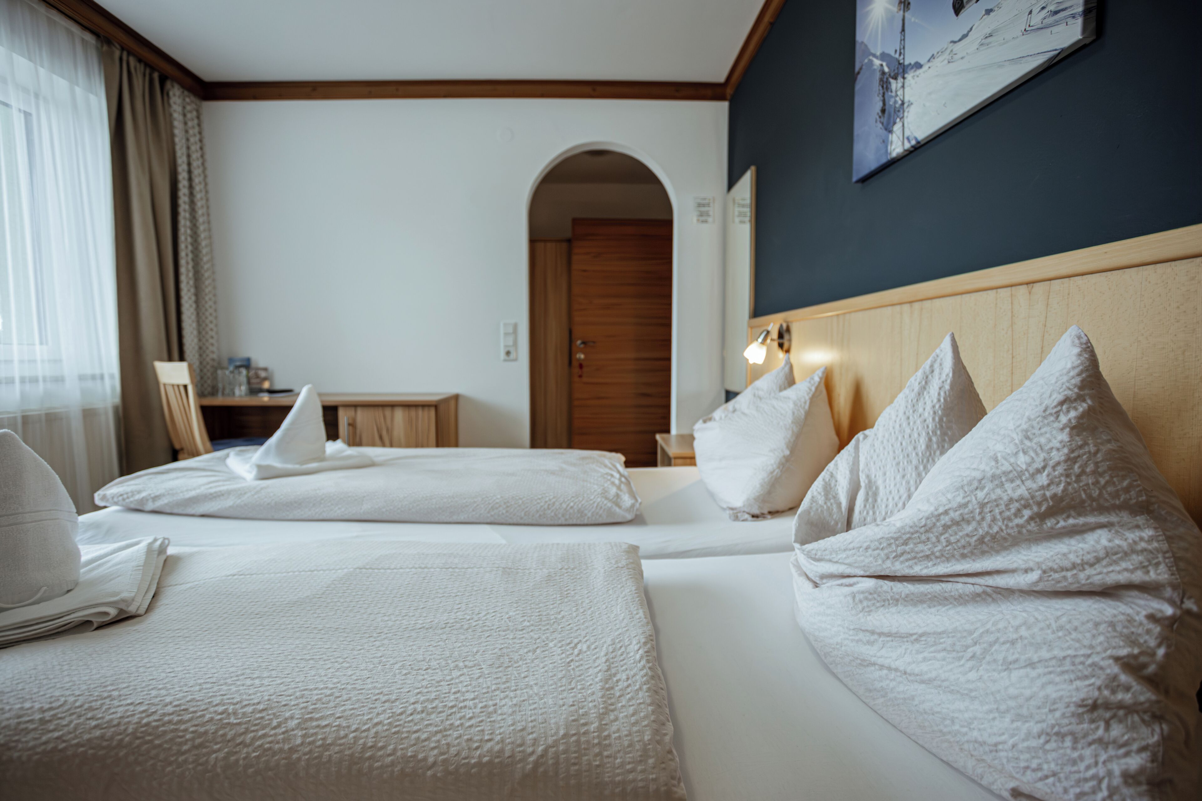 A hotel room with two beds, a wooden headboard, a painting on the wall, and a window with curtains.