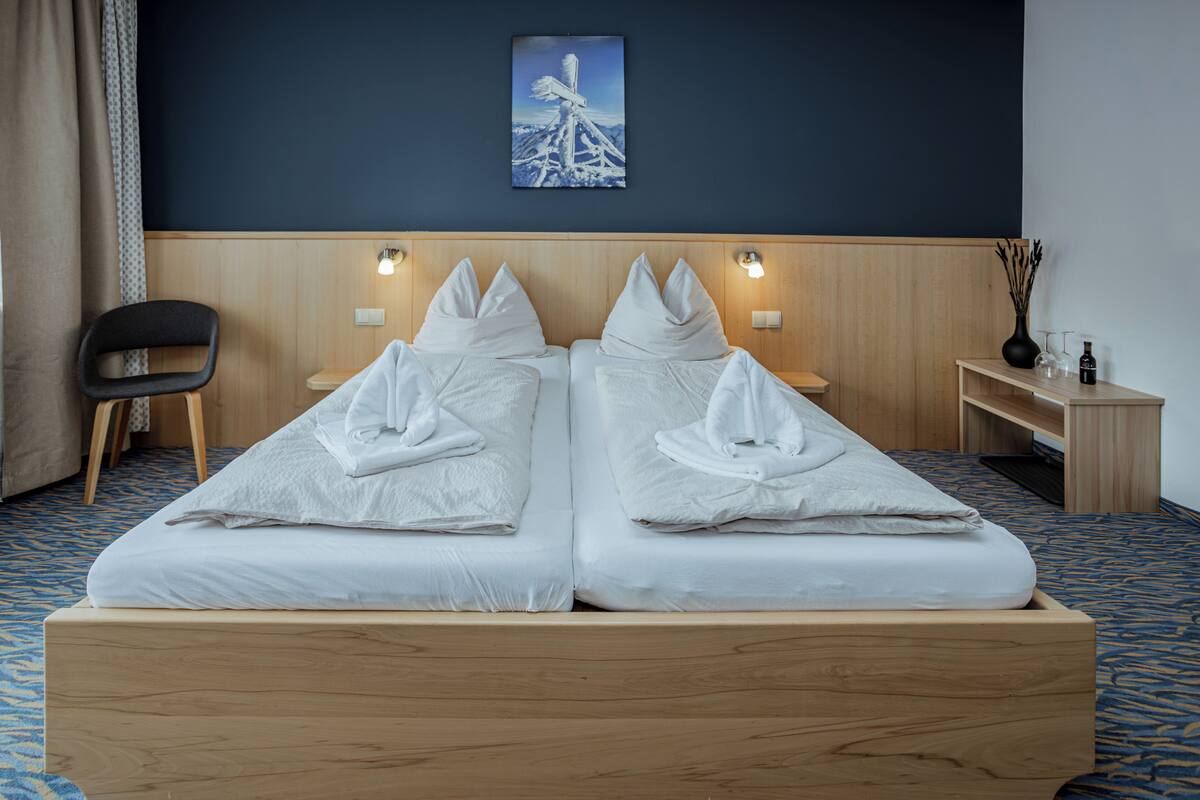 Una cama doble con sábanas y almohadas blancas, cabecera de madera, mesita de noche con lámpara y un cuadro en la pared.