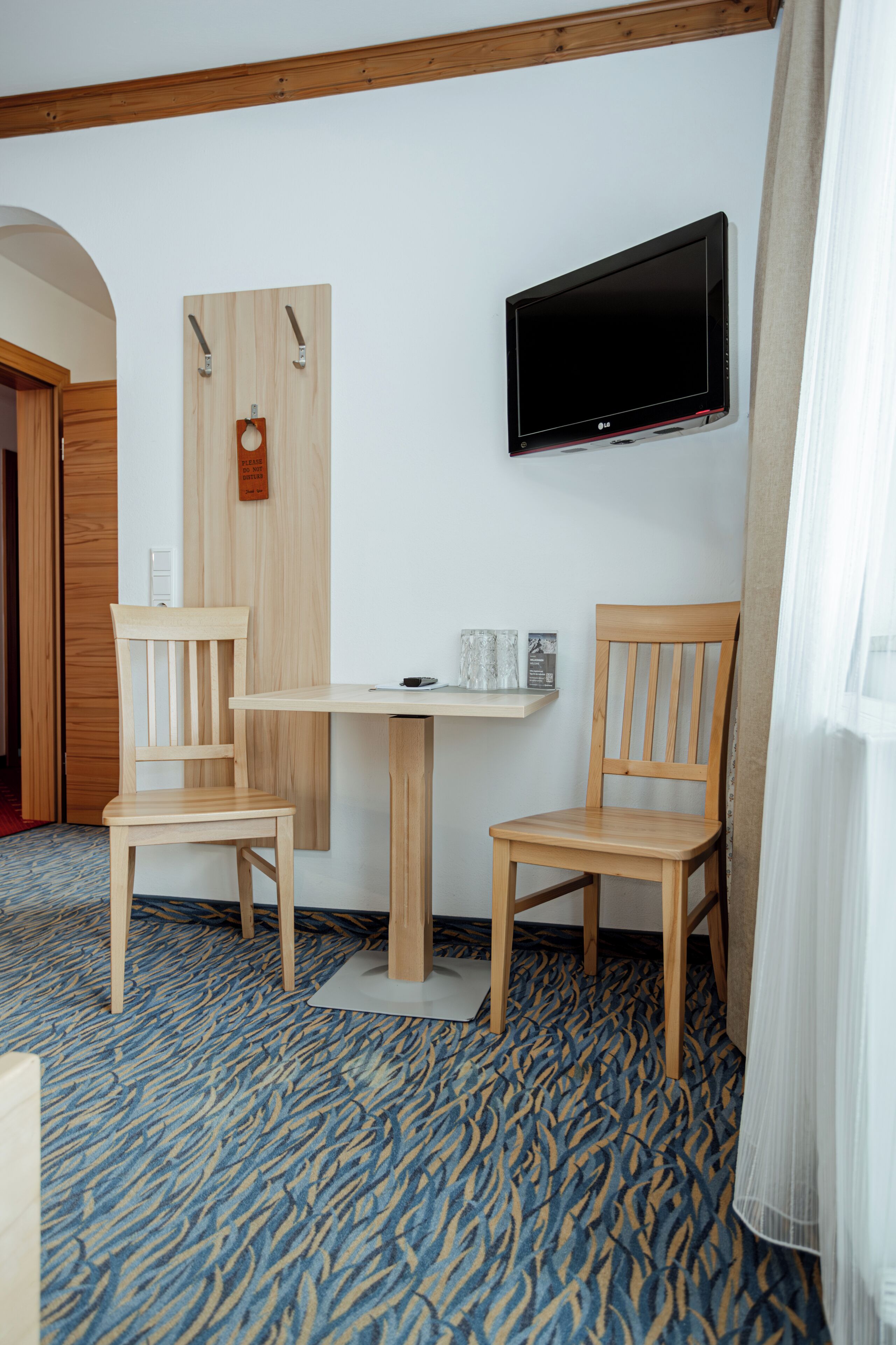 Habitación de hotel con dos sillas de madera, una mesita, televisión montada y alfombra estampada.