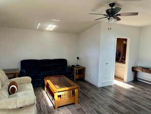 Un soggiorno con un divano blu scuro, un tavolino in legno, una poltrona beige, una ventola a soffitto e una cucina visibile attraverso un'apertura.