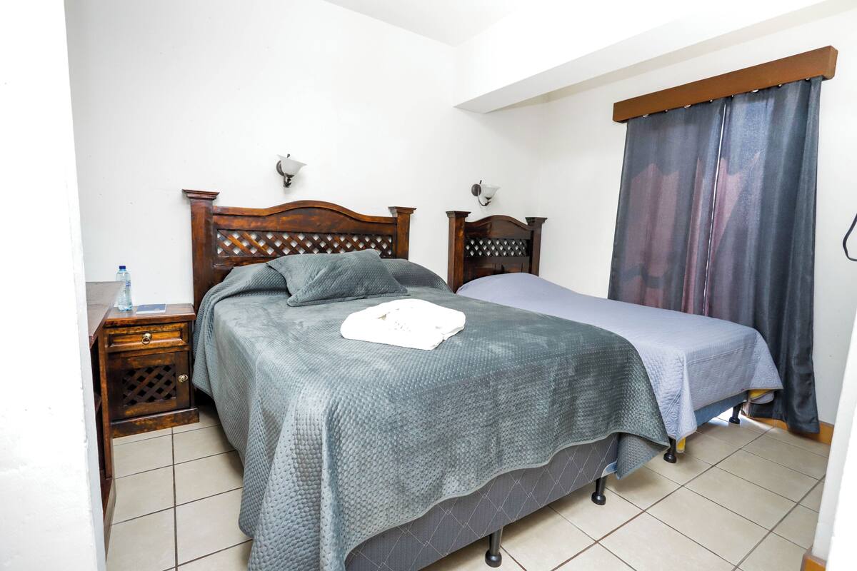 Una camera da letto con una testiera in legno, un letto con copripiumino grigio, un comodino con una bottiglia d'acqua e una tenda alla finestra.
