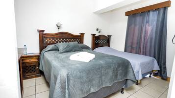 A bedroom with a wooden headboard, a bed with a grey bedspread, a bedside table with a water bottle, and a curtain on the window.