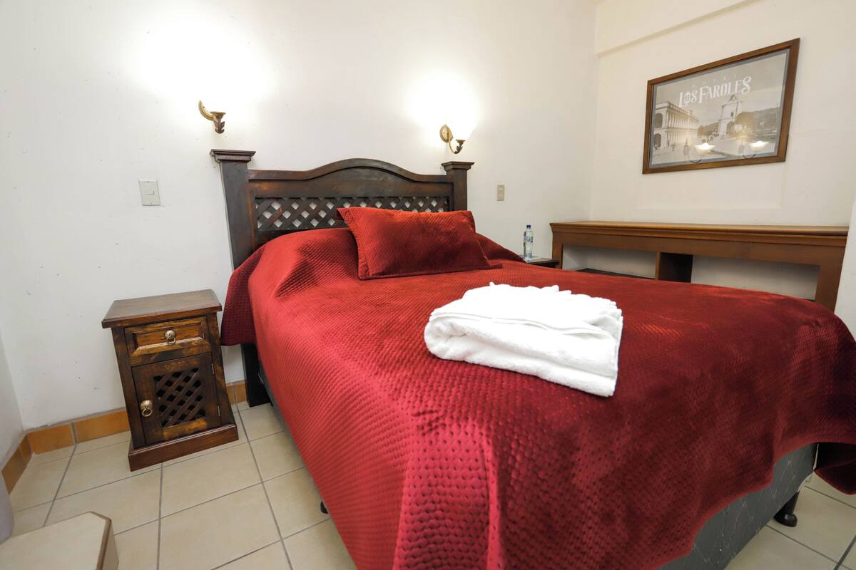 Un letto rifatto con una coperta rossa e cuscini, un comodino in legno con un cassetto e un quadro appeso al muro.