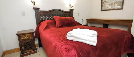 A neatly made bed with a red blanket and pillows, a wooden nightstand with a drawer, and a framed picture on the wall.
