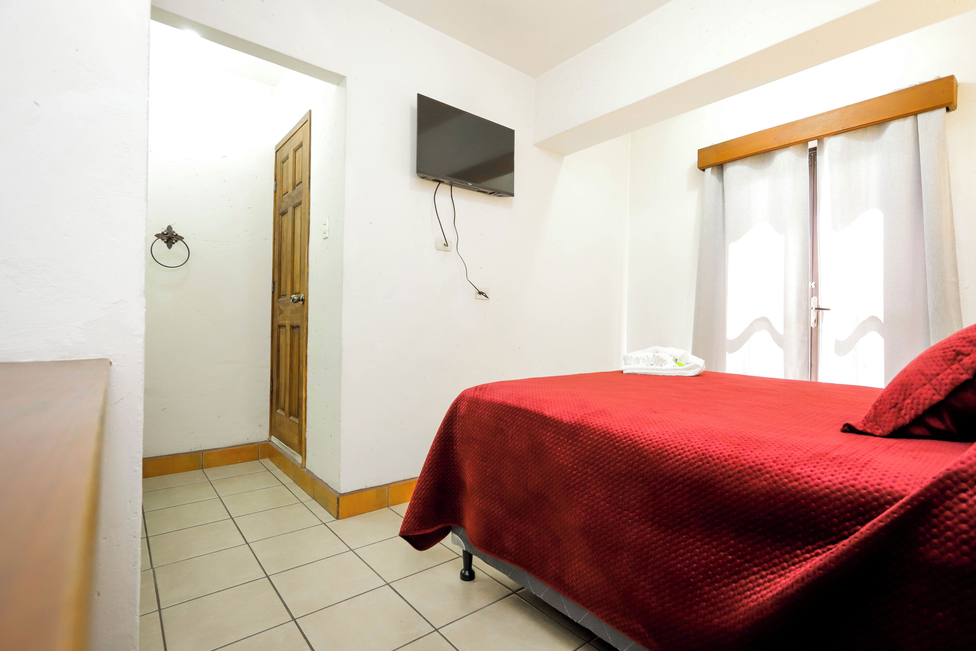 Ein Schlafzimmer mit Bett, Wandfernseher, Tür und einem mit Vorhängen versehenen Fenster.