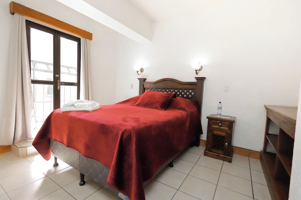 Un letto rifatto con un copriletto rosso, una testiera in legno e due lampade fissate al muro.