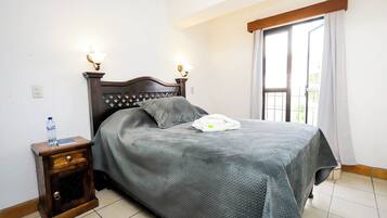 Een netjes opgemaakt bed met een grijze spreidek, een houten nachtkastje met een fles en een boek, en een raam met gordijnen.