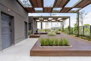 An outdoor area with a wooden deck, planters with green plants, a covered walkway, and a building with grey shutters. - Modern and Sophisticated Studio - Santo Amaro | Near US Consulate and Metro (Santo Amaro São Paulo)