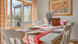 A dining area with a table set for four, white chairs, a red and white checkered table runner, a vase with dried flowers, and a framed floral painting on the wall.