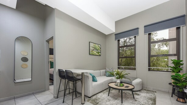 Una sala de estar moderna con un sofá blanco, una mesa de centro redonda y un comedor con una mesa pequeña y sillas. Hay ventanas grandes con persianas, un espejo en la pared y una planta en maceta.