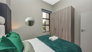 Un dormitorio moderno con una cama, almohadas verdes, una manta azul verdoso, un espejo, un armario y una ventana.