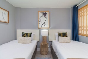 Une chambre d’hôtel avec deux lits, une lampe de sol en rotin, une œuvre d’art accrochée au mur et une fenêtre ornée de rideaux à motifs.