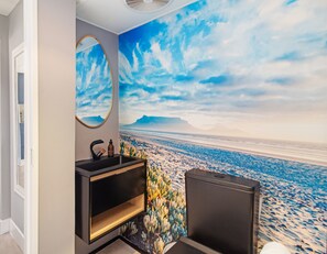 A bathroom with a large wall mural of a beach scene, a round mirror, and a modern sink. - Beachfront Bliss with Pool & Ocean Views (Cape Town)