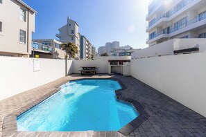 Une piscine dotée d’une terrasse carrelée, entourée d’immeubles avec des balcons, sous un ciel dégagé.