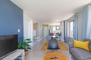 A modern living room with a flat-screen TV, a sofa with a yellow pillow, a dining area with chairs, a kitchen in the background, and a large window with curtains.