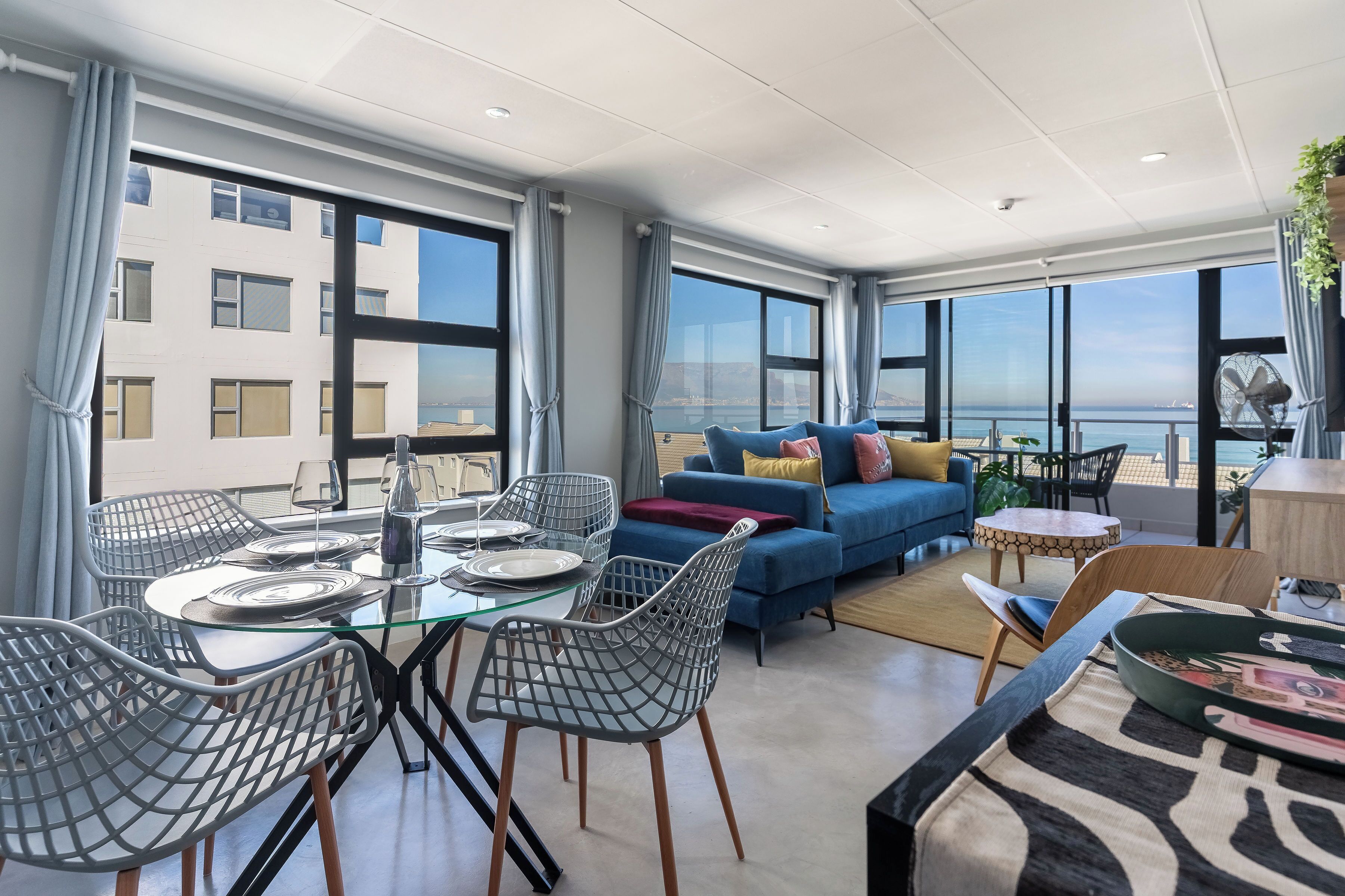 A modern living room with a glass dining table, grey wireframe chairs, a blue sofa with cushions, and large windows offering a view of the sea.