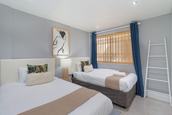 Une chambre d’hôtel avec deux lits, une échelle, une fenêtre avec des rideaux, une œuvre d’art accrochée au mur et une lampe de chevet.