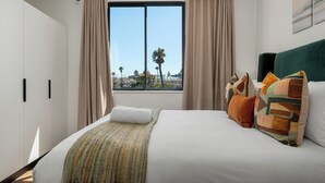 Hotelkamer met een groot bed, beige gordijnen, uitzicht op palmbomen en een stadsbeeld.