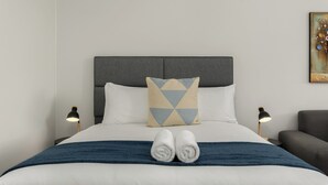 A neatly made bed with two pillows, a blue blanket, and two rolled towels. There are two bedside lamps and a chair to the right. - On Park | Modern Century City Studio (Cape Town)