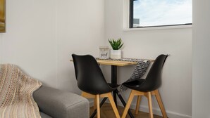 A small dining area with a table, two black chairs, a grey sofa, and a window with a view of the sky. - On Park | Modern Century City Studio (Cape Town)