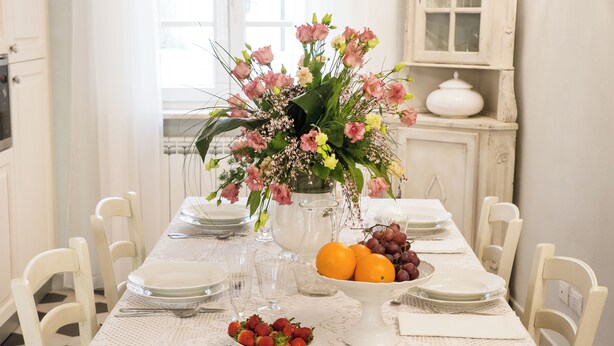 A dining table set with plates, cutlery, a vase of flowers, and a bowl of fruit.