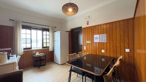 Une pièce avec un mur lambrissé en bois, une table à manger avec des chaises, un réfrigérateur, une petite table avec une bouteille d’eau et une fenêtre avec des rideaux.