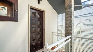 Une porte en bois sombre ornée d’un motif géométrique, une fenêtre à cadre blanc avec des stores horizontaux et un pilier en pierre.