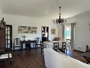 A spacious living room with a white sofa, a dining area with a table and chairs, a chandelier, and a view through a window.