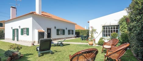 A white house with a red-tiled roof, a chimney, and a small garden with wicker chairs and a grill.