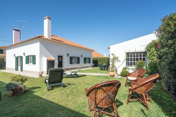 A white house with a red-tiled roof, a chimney, and a small garden with wicker chairs and a grill.