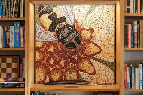 A framed mosaic artwork with a central figure surrounded by a patterned border, displayed on a shelf with books.