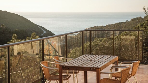 A balcony with a wooden table and chairs, overlooking a coastal landscape with hills and the sea. - Magoito • Refuge for nature lovers (São João das Lampas)