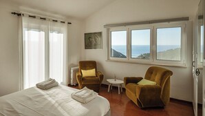 A hotel room with a bed, two armchairs, a small table, and a view of the sea through large windows. - Magoito • Refuge for nature lovers (São João das Lampas)