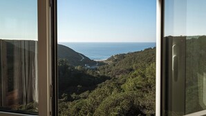 A view from a window overlooking a coastal landscape with a road leading to a beach, surrounded by dense greenery. - Magoito • Refuge for nature lovers (São João das Lampas)