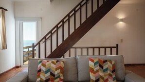 A living room with a sofa, two colorful pillows, a wooden staircase, and a view through a glass door. - Magoito • Refuge for nature lovers (São João das Lampas)