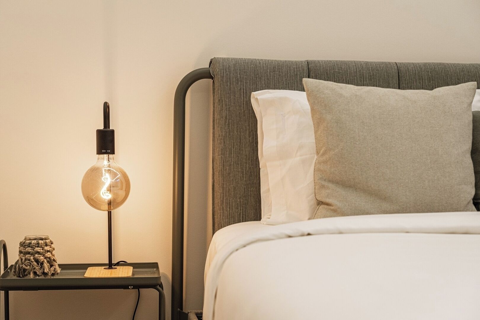 Een netjes opgemaakt bed met witte lakens en twee kussens, een bedlamp met een glazen kap en een klein houten dienblad met een potje en een kaars.
