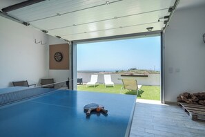 Une table de ping-pong avec un filet, un jeu de fléchettes et deux chaises sur un balcon offrant une vue dégagée sur l’océan.