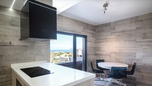 Une cuisine moderne dotée d’un grand îlot central, d’un coin repas avec une table ronde et des chaises, et offrant une vue sur la mer grâce à de grandes fenêtres.