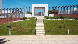 A modern building with a central staircase leading to a glass door entrance, surrounded by a symmetrical garden and a metal fence.