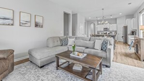A modern living room with a sectional sofa, a coffee table, and a rug. There is a kitchen in the background with a dining area.