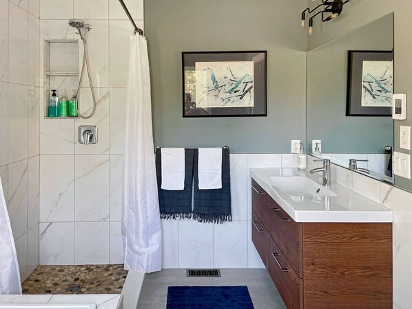 A modern bathroom with a shower, a sink, and a mirror. There are towels hanging on the wall and a small shelf with toiletries.
