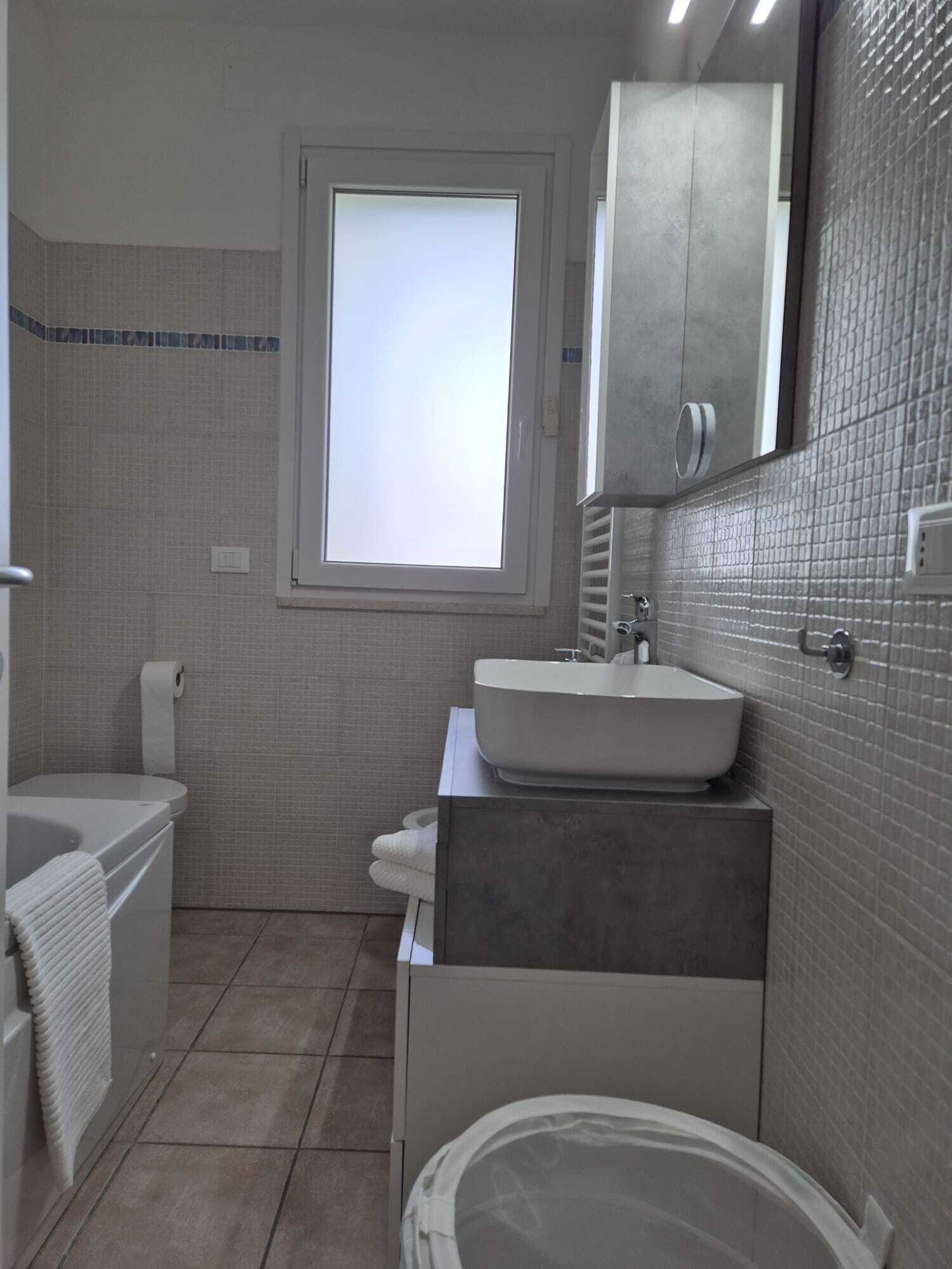 Een moderne badkamer met een badkuip, wastafel en toilet. Een raam zorgt voor natuurlijk licht, en boven de wastafel hangt een spiegel.