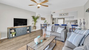 A modern living room with a grey sofa, a coffee table, and a television. There is a ceiling fan and a kitchen area in the background. - Ocean view condo in Surf City! (Surf City)