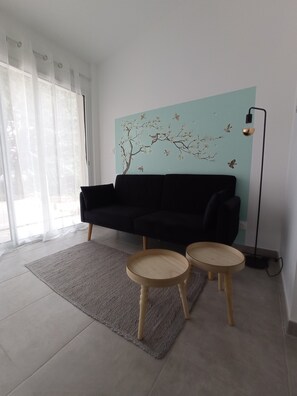 A modern living room with a black sofa, two wooden side tables, a light blue wall with a tree mural, and a floor lamp.