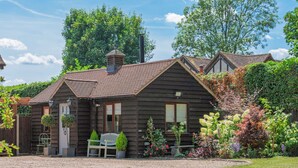 A wooden cabin with a gravel driveway, a bench, and a small garden with various plants and flowers. - Rose Cottage - UK49048 (Banstead)