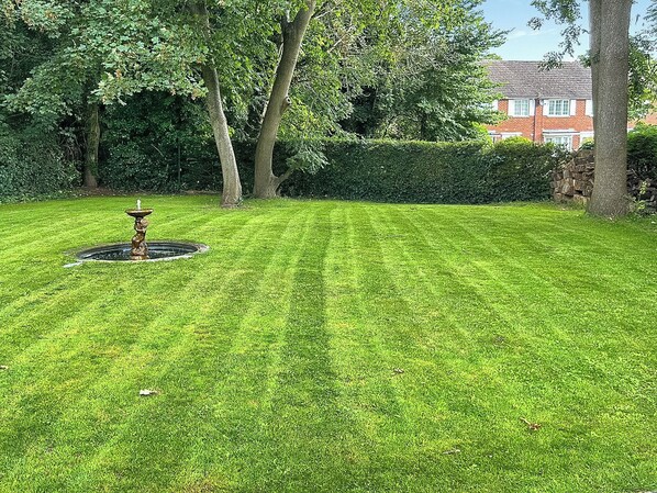 A well-maintained lawn with a central fountain, surrounded by mature trees and a hedge.