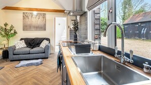 A modern kitchen with a large window, wooden ceiling beams, and a grey sofa in the living area. - Rose Cottage - UK49048 (Banstead)